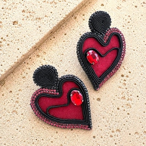 Puede incluir: Un par de pendientes en forma de corazón con un centro de terciopelo rojo intenso, rodeado de detalles de cordón negro y un borde de pequeños cristales rojos. Cada pendiente incluye un poste de cordón negro y un cristal rojo ovalado.