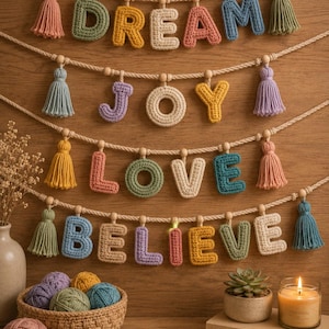 Op de afbeelding: Een wandhanger met vier horizontale touwen waaraan gehaakte letters hangen die de woorden DREAM, JOY, LOVE en BELIEVE vormen. De letters zijn in diverse pastelkleuren en worden afgewisseld met houten kralen en hangende garenkwastjes.