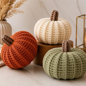 May include: Three crocheted pumpkins in orange, cream, and sage green. Each pumpkin features a ribbed texture and a small, brown, textured stem at the top. The pumpkins are round and display a consistent, handmade yarn stitch pattern throughout.
