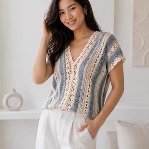Puede incluir: Una blusa de ganchillo de manga corta con un patrón de rayas verticales en tonos alternos de azul polvoriento y crema. La prenda tiene un escote en V con borde festoneado y un diseño de punto calado en toda la tela.