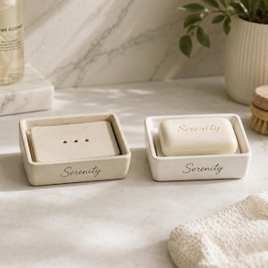 Pode incluir: Dois saboneteiras retangulares bege com a palavra Serenity impressa em letra cursiva preta na frente. Uma contém um inserto bege com três orifícios de drenagem, enquanto a outra segura uma barra de sabão lisa com a inscrição Serenity gravada.