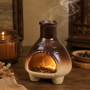 May include: A brown ceramic incense burner shaped like a small chiminea with a wide base, three short legs, and a chimney top. The burner features a gradient glaze from dark brown at the top to a lighter beige at the base, with smoke rising.