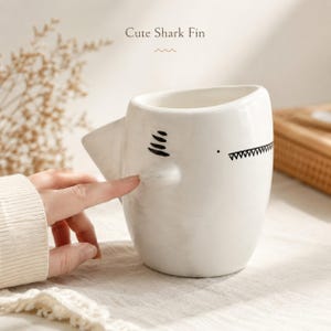 May include: A white ceramic mug shaped like a shark, featuring a protruding fin on the side, a black painted eye, and a jagged shark tooth mouth design. The text "Cute Shark Fin" is printed above the mug in a simple, dark brown font.