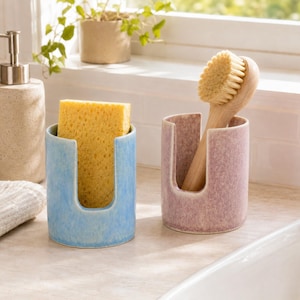 May include: A light blue ceramic holder containing a yellow rectangular sponge and a mauve ceramic holder containing a wooden dish brush with light tan bristles. Both holders feature a U-shaped cutout on the front side for easy access.
