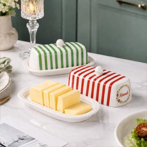 Könnte beinhalten: Zwei Butterdosen aus Keramik mit Deckeln. Ein Deckel hat grüne Streifen, der andere rote Streifen und ein florales Kranzmotiv mit dem Namen Hannah in der Mitte. Ein Stück gelbe Butter liegt in Scheiben auf dem Teller der roten Butterdose.