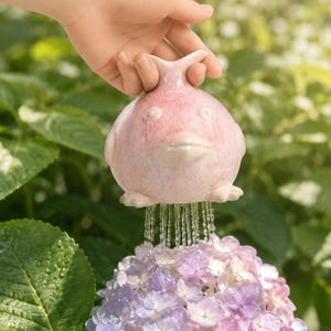 May include: A light pink ceramic watering tool shaped like a round fish with a wide mouth and fins. The object features a perforated base that releases a steady stream of water onto a cluster of purple and white hydrangea flowers below.