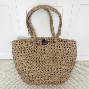 Può includere: Una borsa tote beige realizzata in materiale intrecciato e testurizzato. La borsa presenta due spallacci intrecciati abbinati e un dettaglio a linguetta rettangolare marrone scuro visibile vicino all'apertura centrale superiore.