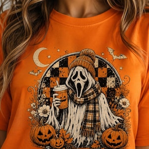 May include: An orange t-shirt featuring a graphic print of a ghost character wearing a beanie and scarf, holding a pumpkin-faced coffee cup. The design includes a checkered background, two jack-o'-lanterns, autumn leaves, bats, and a crescent moon.
