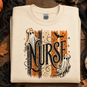 May include: A cream-coloured crewneck sweatshirt featuring a graphic print with the word NURSE in large black lettering. The design includes two ghosts wearing nurse caps, orange and black vertical brush strokes, bats, and small stars.