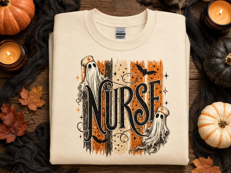May include: A cream-coloured crewneck sweatshirt featuring a graphic print with the word NURSE in large black lettering. The design includes two ghosts wearing nurse caps, orange and black vertical brush strokes, bats, and small stars.
