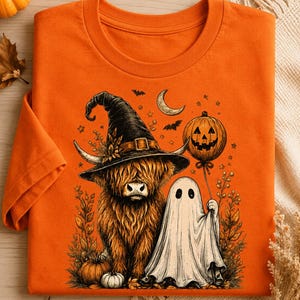 May include: An orange t-shirt featuring a graphic illustration of a Highland cow wearing a black witch hat, standing next to a white ghost holding a pumpkin balloon. The design includes small bats, a crescent moon, and autumn foliage elements.