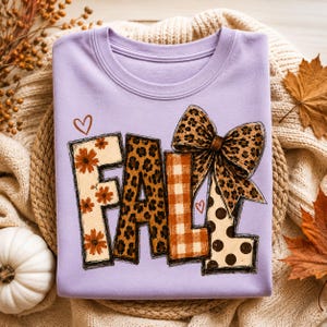 May include: A light purple crewneck sweatshirt featuring the word FALL in large block letters. Each letter has a different pattern: floral, leopard print, orange plaid, and brown polka dots. A large leopard print bow sits atop the letter L.