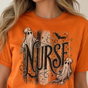 May include: An orange short-sleeved t-shirt featuring a graphic print with the word NURSE in large black letters. The design includes two white ghosts wearing nurse caps, small black bats, and vertical distressed stripes in black and tan.