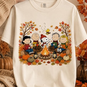 Puede incluir: Una camiseta de manga corta color crema con un gráfico de los personajes de Peanuts sentados en troncos alrededor de una fogata. El diseño incluye hojas de otoño, calabazas, linternas y los personajes asando malvaviscos.