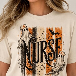 May include: A cream-coloured t-shirt featuring a graphic print with the word NURSE in large black lettering. The design includes two ghosts wearing nurse caps, black bats, and vertical stripes in shades of orange, black, and beige.