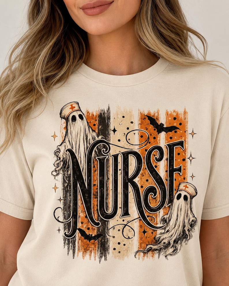 May include: A cream-coloured t-shirt featuring a graphic print with the word NURSE in large black lettering. The design includes two ghosts wearing nurse caps, black bats, and vertical stripes in shades of orange, black, and beige.