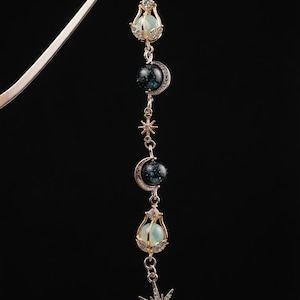 Puede incluir: Un marcapáginas de metal plateado con una cadena colgante de dijes de temática celestial. El diseño incluye dos cuentas esféricas de color azul pálido en engastes dorados, dos esferas de purpurina azul oscuro con lunas crecientes plateadas y estrellas.