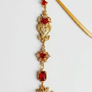 Puede incluir: Un marcapáginas de metal dorado con un diseño de dijes colgantes. El adorno incluye un pequeño cristal rojo, una pieza de filigrana en forma de corazón con una rosa roja, un marco cuadrado con una piedra roja y detalles claros, y una rosa roja con corona.