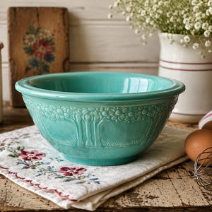 May include: A turquoise ceramic mixing bowl featuring a raised floral and tree pattern around the exterior. The bowl has a smooth, glossy finish and a wide, rounded rim, sitting on a white cloth with a small red and green floral embroidery.