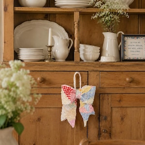 May include: A fabric bow ornament featuring a patchwork design of various colourful patterned textiles, including red polka dots, blue florals, and cream-coloured fabrics. The bow has a braided fabric loop at the top for hanging purposes.