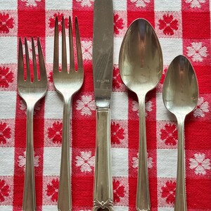 May include: A five-piece stainless steel flatware set consisting of two forks, one dinner knife, one tablespoon, and one teaspoon. Each piece features a decorative floral motif at the base of the handle, with the knife blade marked as stainless.