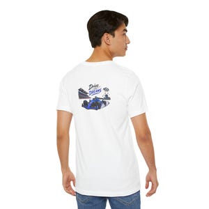 Puede incluir: Una camiseta blanca de manga corta con un gráfico en la parte posterior. El diseño muestra un coche de carreras azul en una pista con una bandera a cuadros y el texto Drive Your Dreams. La camiseta se combina con pantalones vaqueros azules.