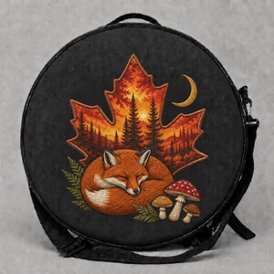 May include: A round black bag featuring an embroidered design of a sleeping orange fox curled up next to three mushrooms. The scene is set against a large maple leaf containing a forest landscape with pine trees, a crescent moon, and a sun.