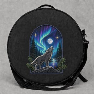 Könnte beinhalten: Eine schwarze, runde Umhängetasche mit einem zentralen Grafikdruck eines grauen Wolfes, der den Vollmond anheult. Die Szene zeigt einen dunklen Wald, ein reflektierendes Gewässer und leuchtend violette sowie grüne Polarlichter am Himmel.