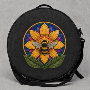 Könnte beinhalten: Eine runde schwarze Tasche mit einem zentralen Stickmotiv einer gelben Sonnenblume und einer Biene in der Mitte. Die Blume ist von einem farbenfrohen geometrischen Mandalamuster in Blau-, Violett- und Orangetönen umgeben, mit zwei grünen Blättern.
