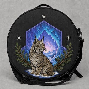 May include: A round black crossbody bag featuring a central illustration of a lynx sitting in a snowy landscape. The lynx is framed by a blue hexagonal archway with icicles, flanked by green pine branches and white stars on a dark background.