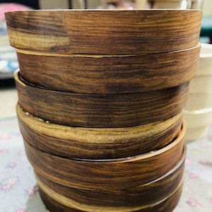 Op de afbeelding: Een stapel ronde houten kommen met een rijk, donkerbruin nerfpatroon met lichtere goudbruine strepen. De kommen hebben gladde, afgeronde randen en een consistente cilindrische vorm, die de natuurlijke textuur van de houtnerf laat zien.