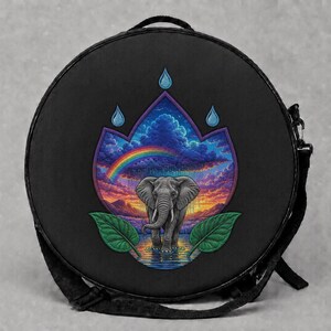 May include: A round black crossbody bag featuring a central graphic print of a gray elephant standing in water. The scene includes a vibrant rainbow, a sunset sky with clouds, and two green leaves framing the base, topped by three blue raindrops.