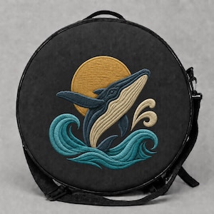May include: A round black crossbody bag featuring an embroidered design of a blue whale leaping from teal ocean waves toward a golden yellow sun. The bag has a top handle, a black adjustable strap, and a zipper closure along the outer edge.
