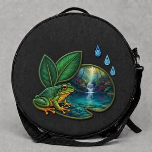 Könnte beinhalten: Eine runde schwarze Umhängetasche mit einem farbenfrohen Grafikdruck eines grünen Frosches, der auf einem Seerosenblatt neben einem Wasserfall in einem Wald sitzt. Das Design zeigt zwei große grüne Blätter und drei kleine blaue Wassertropfen.