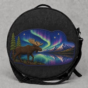 May include: A round black bag featuring a printed graphic of a brown moose standing near a lake with pine trees, snow-capped mountains, and a vibrant green and purple aurora borealis in the night sky with visible stars above the landscape.
