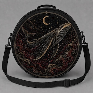 May include: A round black crossbody bag featuring a detailed illustration of a humpback whale swimming through dark, swirling clouds and waves under a golden crescent moon and stars. The bag has a top handle and an adjustable black shoulder strap.