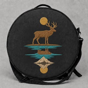 May include: A round black bag featuring an embroidered design of a brown stag standing on a small island beneath a gold sun. Below the island, a teal water reflection and a mountain peak are mirrored, ending with a second gold sun at the bottom.