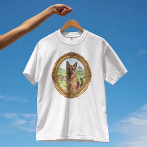 May include: A white short-sleeved t-shirt featuring a central graphic of a German Shepherd dog sitting within an ornate, oval-shaped gold frame. The background inside the frame depicts a landscape with a small village and a castle.