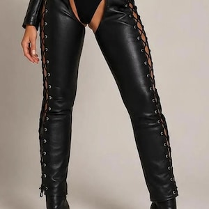 May include: Black leather chaps featuring a high-waisted belt with silver-toned metal hardware. The legs include full-length side lacing with silver eyelets and black cord ties. The design is open at the front and back, paired with black boots.