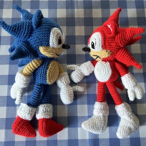 May include: Two handmade crochet plush toys representing stylised characters. One figure is blue with tan accents and red shoes, while the other is red with tan accents and white shoes. Both feature white gloves, large eyes, and spiky hair designs.