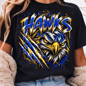 May include: A black t-shirt featuring a graphic print of a hawk head with yellow eyes and a yellow beak. The word HAWKS is printed in large, stylized blue and yellow letters above the bird. The design includes blue and yellow claw marks.