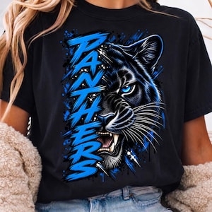 May include: A black t-shirt featuring a graphic print of a panther's face on the right side with bright blue eyes and bared teeth. The word PANTHERS is written vertically on the left in a stylized, brush-stroke blue font with blue paint splatters.