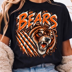 Può includere: Una t-shirt nera con una stampa grafica di un orso bruno ringhiante con occhi arancioni. Sopra l'orso, la parola BEARS è scritta in un carattere arancione usurato e gocciolante. Tre segni di artigli diagonali tagliano il design in arancione.