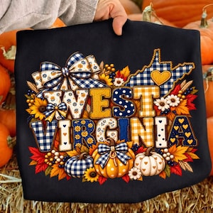 Può includere: Una t-shirt nera con la scritta WEST VIRGINIA in stile patchwork con motivi a quadretti blu e bianchi, pois gialli e fiori. Il design include una mappa della Virginia Occidentale, zucche, girasoli e foglie autunnali.
