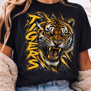 Puede incluir: Una camiseta negra con un gráfico de una cabeza de tigre rugiendo con detalles de pelaje amarillo y blanco. La palabra TIGERS está impresa verticalmente en una fuente amarilla, negrita y desgastada, acentuada por rayas amarillas como garras.