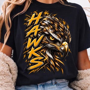 May include: A black short-sleeved t-shirt featuring a graphic of a hawk head in profile with yellow and white feathers. The word HAWKS is printed vertically in a bold, distressed yellow font to the left of the bird, accented by yellow paint splatters.