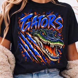 May include: A black t-shirt featuring a graphic of an alligator head with an open mouth and sharp teeth. Above the alligator, the word GATORS is printed in a stylized blue font. The design includes orange and blue paint splatter effects.