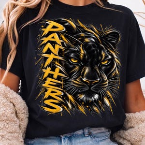 May include: A black short-sleeve t-shirt featuring a graphic print of a panther face with yellow eyes. The word PANTHERS is printed vertically in a distressed yellow font on the left side of the design, accented by yellow paint splatters.
