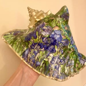 May include: A large seashell featuring a painted floral design with vibrant blue and purple irises set against lush green foliage. The spiral top and the outer rim of the shell are finished with a metallic gold paint, providing a contrast to the detailed botanical artwork covering the main surface.