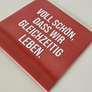Könnte beinhalten: Ein quadratischer roter Untersetzer mit dem weißen Text: VOLL SCHÖN, DASS WIR GLEICHZEITIG LEBEN. Der Untersetzer hat eine glatte, flache Oberfläche und ist schräg positioniert, wobei der deutsche Satz in einer fetten serifenlosen Schrift steht.